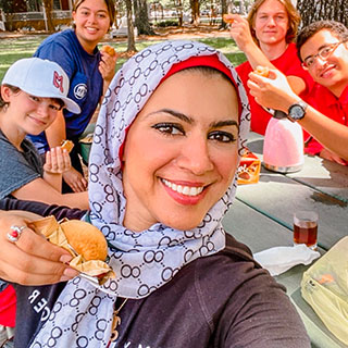 students at picnic table enjoying food while woman holds up some food in front of the camera